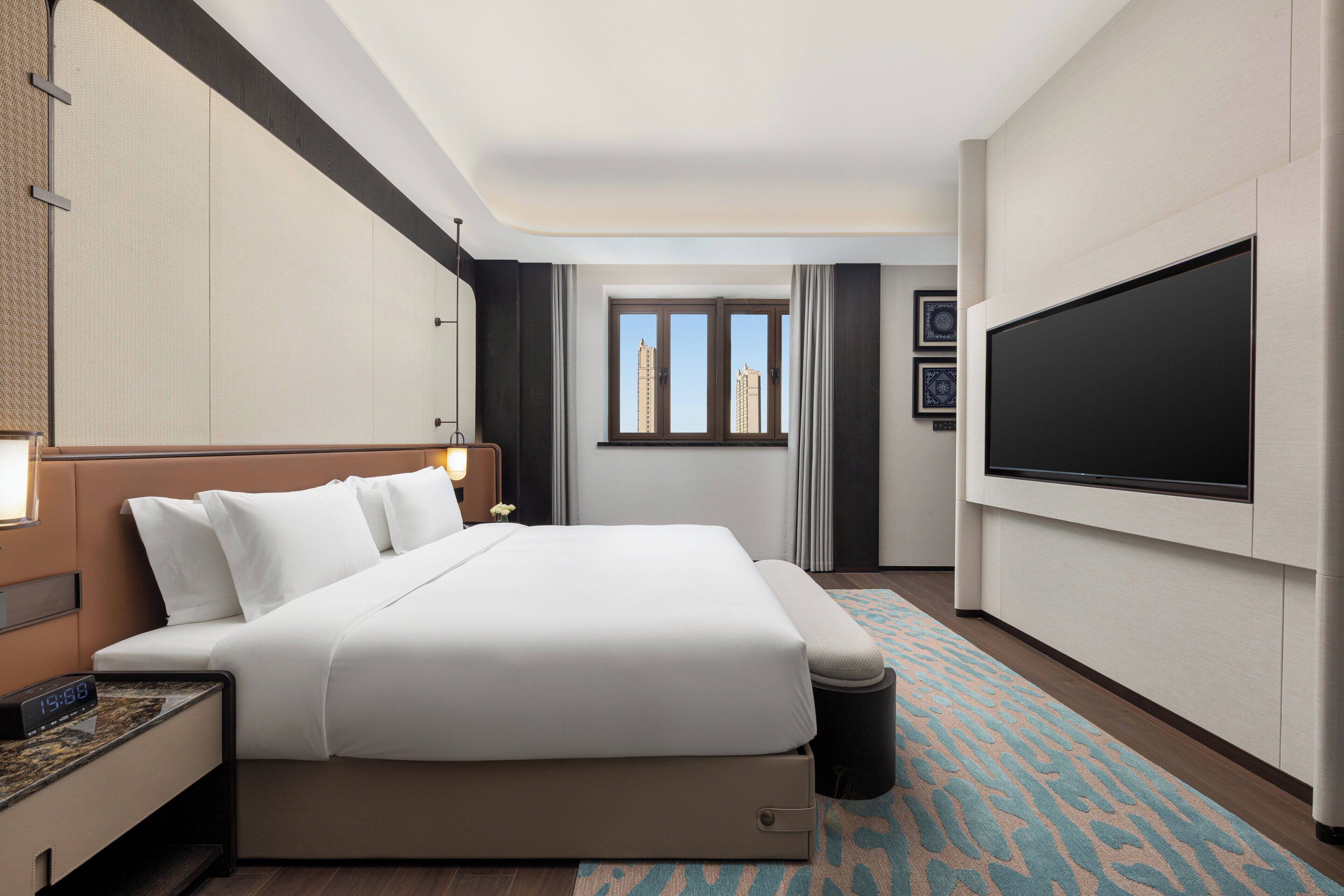 Una habitación de hotel con una cama grande, un televisor de pantalla plana, un ventanal con vistas a edificios y una alfombra estampada.