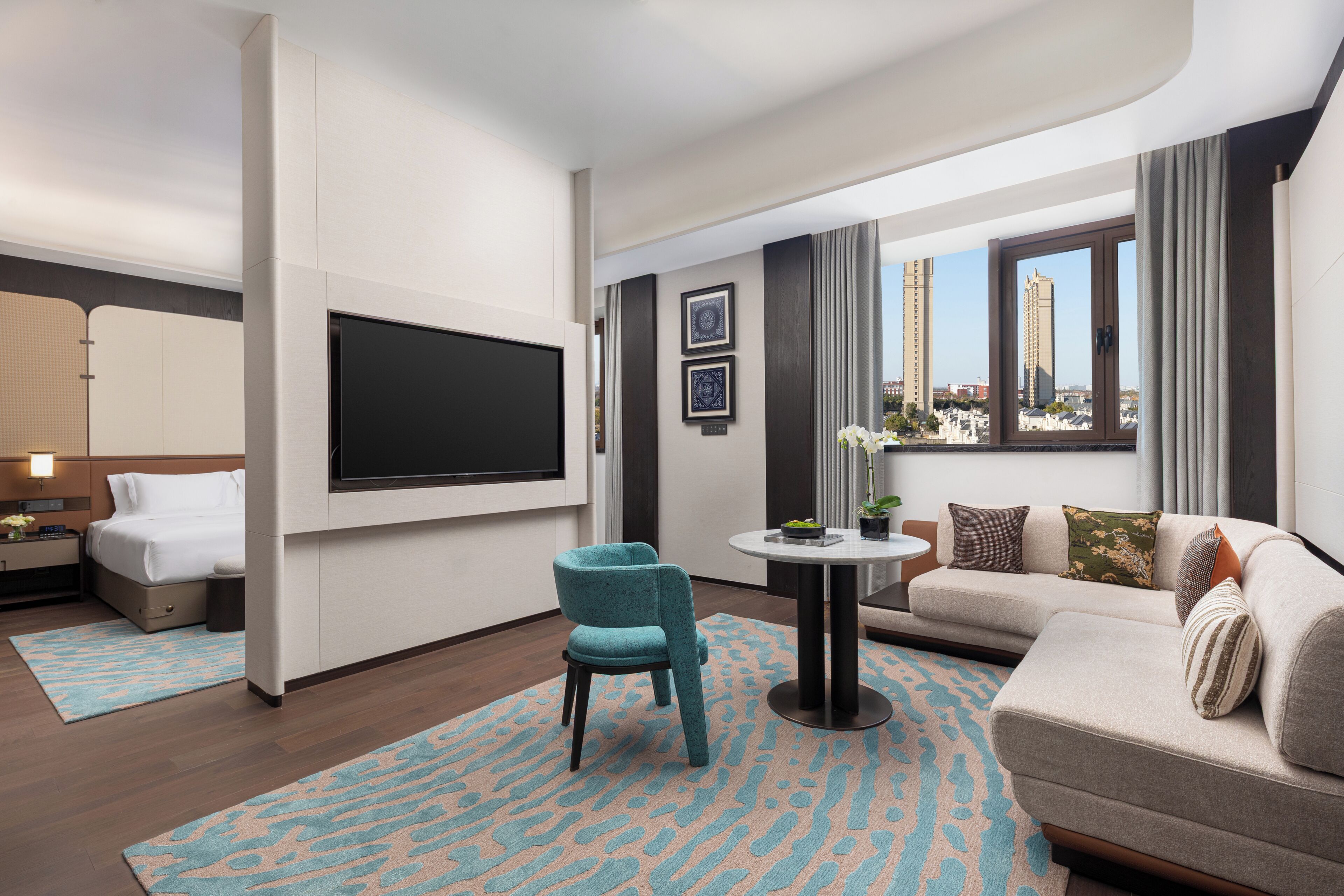 Une chambre d’hôtel moderne dotée d’une télévision à écran plat, d’un canapé, d’un fauteuil, d’une petite table et offrant une vue sur le paysage urbain.