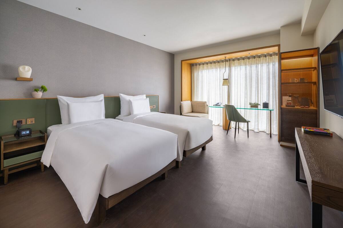 Ein Hotelzimmer mit zwei Betten, einem Schreibtisch, einem Sessel, einem Fernseher und einem Fenster mit Vorhängen.