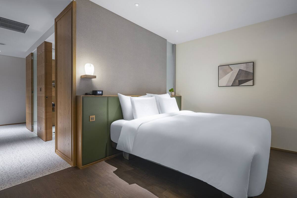 Ein modernes Hotelzimmer mit einem ordentlich bezogenen Bett, einem Nachttisch mit Lampe und einem an der Wand hängenden Kunstwerk.