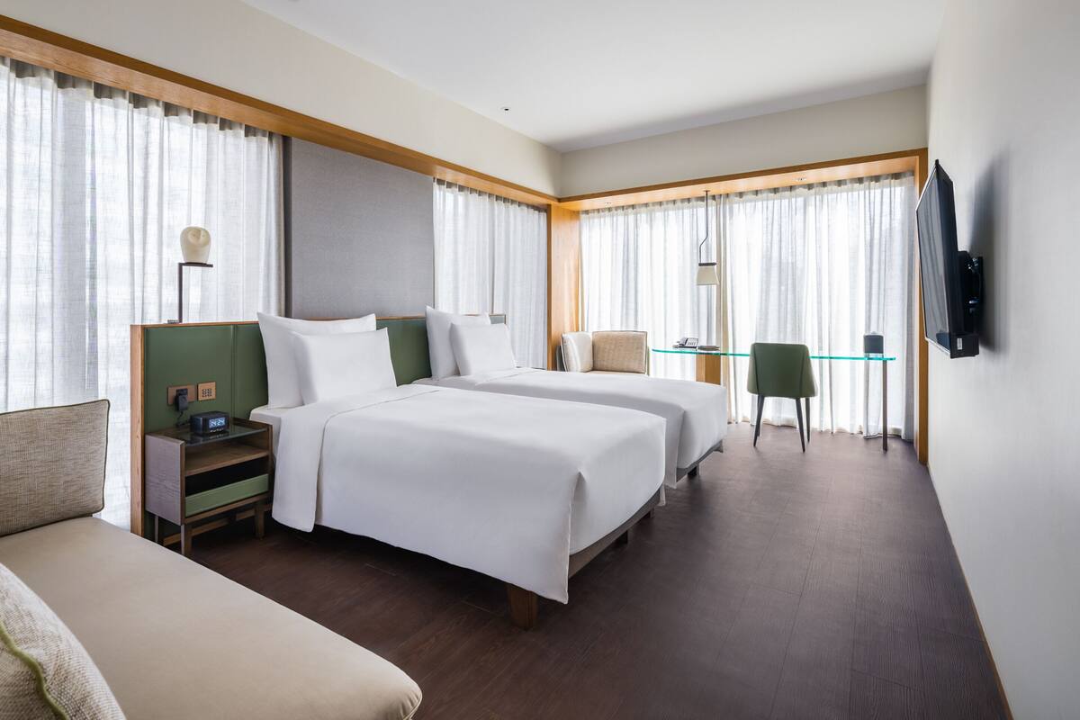 Ein Hotelzimmer mit zwei Betten, einem Schreibtisch, einem Sessel, einem Fernseher und einem Fenster mit Vorhängen.