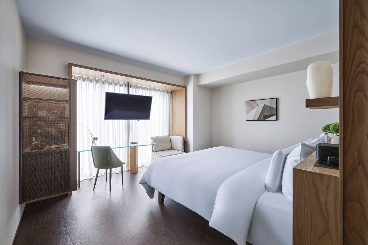 Ein Hotelzimmer mit einem großen Bett, einem Fernseher, einem kleinen Tisch mit Stuhl und einer dekorativen Vase auf einem Regal.