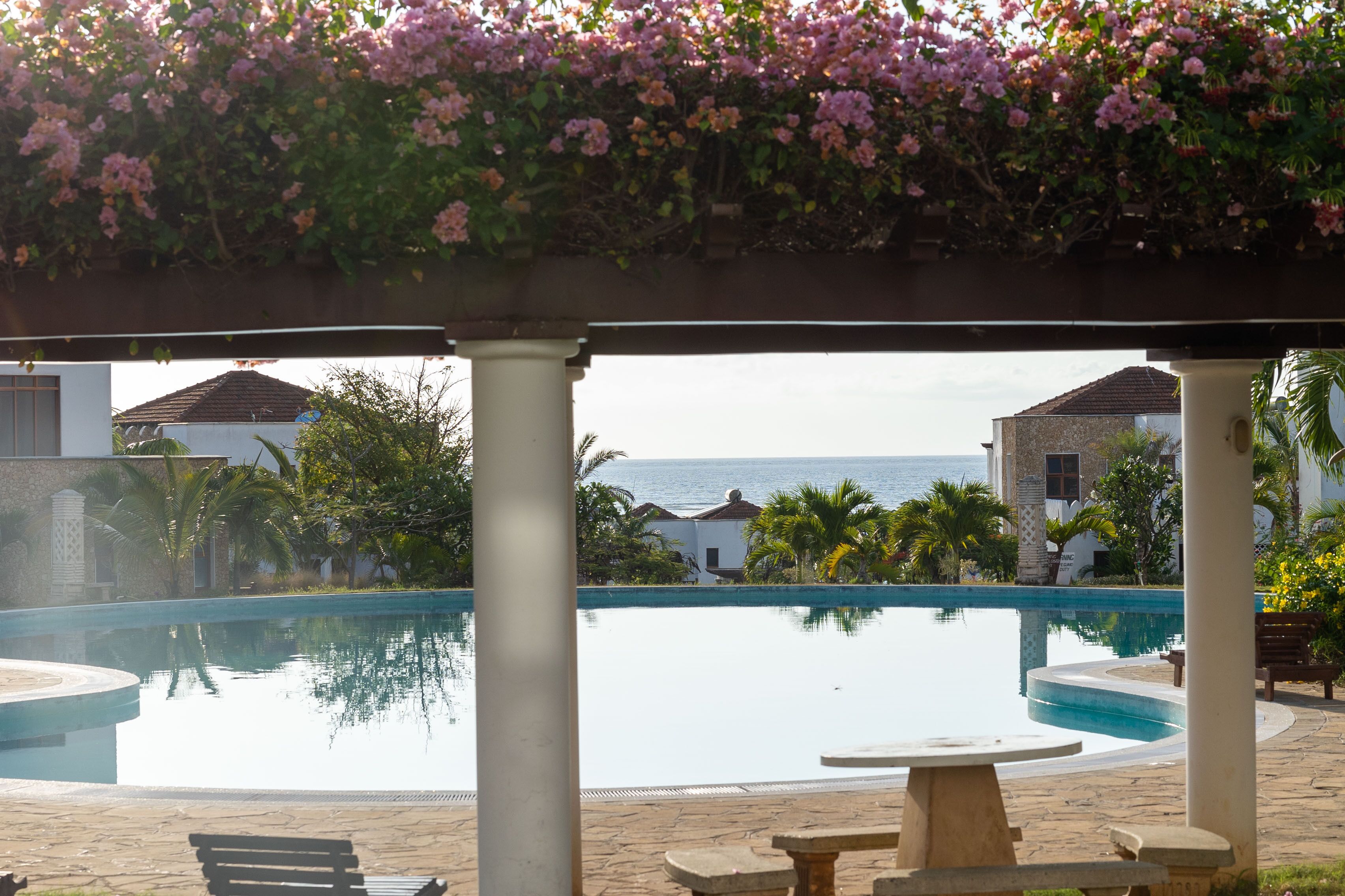 A pool area with a view of the sea, surrounded by columns and a pergola with flowers.