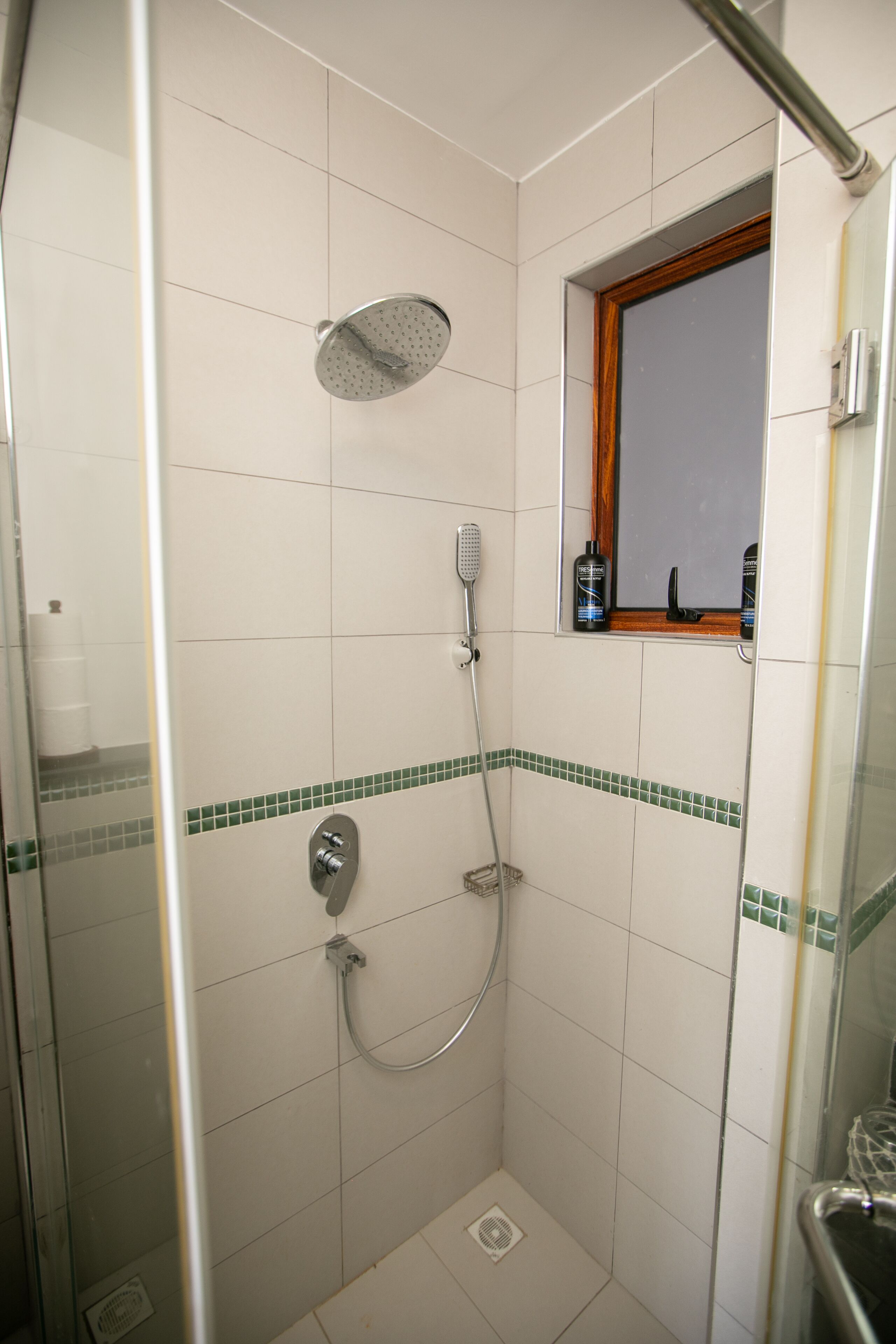 A small, tiled shower area with a handheld showerhead, a wall-mounted faucet, and a window with a wooden frame.