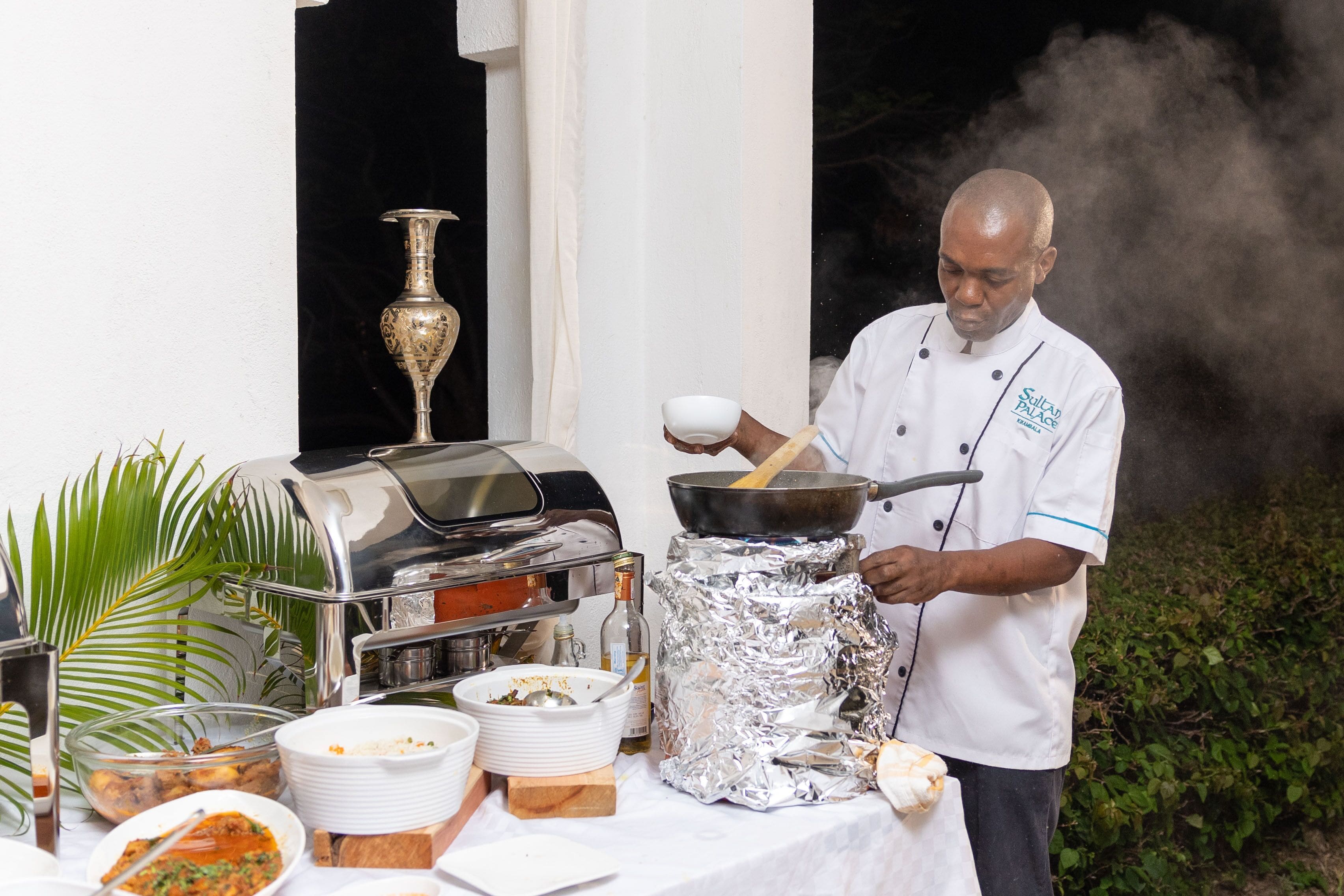 Un chef cocina al aire libre usando una parrilla y varios platos sobre una mesa.