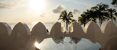 A table with two champagne flutes, a glass of water, and a glass of wine, set against a backdrop of a sunset over the ocean with palm trees.