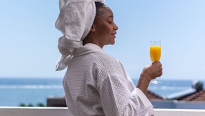 Una persona con bata blanca sosteniendo un vaso de jugo de naranja, de pie en un balcón con vistas despejadas al océano.