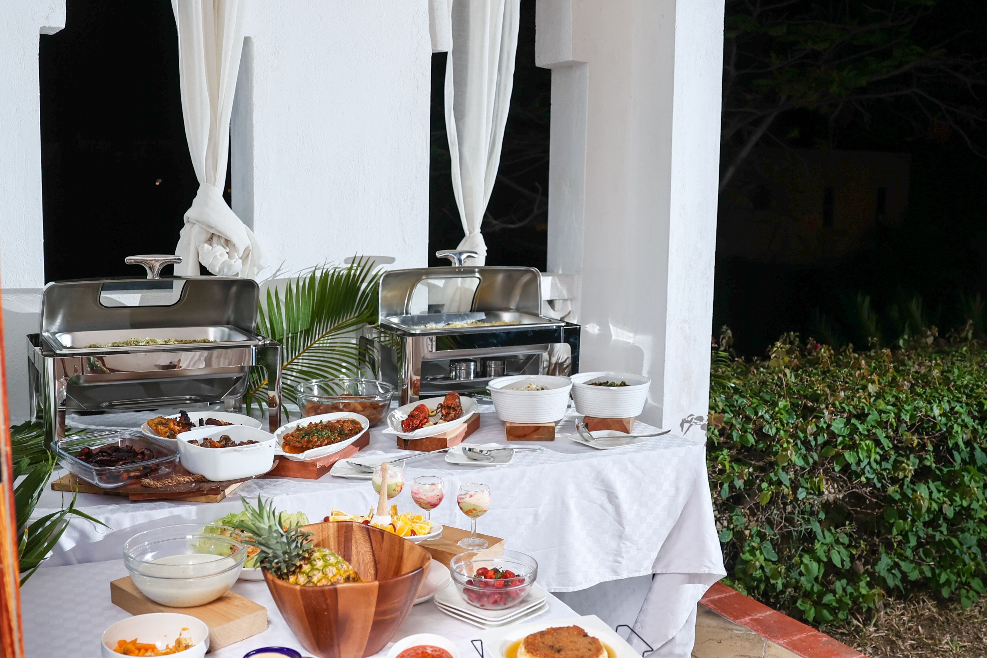 Un bufé al aire libre con distintos alimentos y recipientes sobre una mesa, rodeado de follaje.