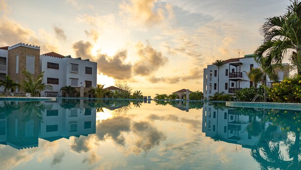 Un resort con piscina cristalina que refleja edificios y un cielo al atardecer.