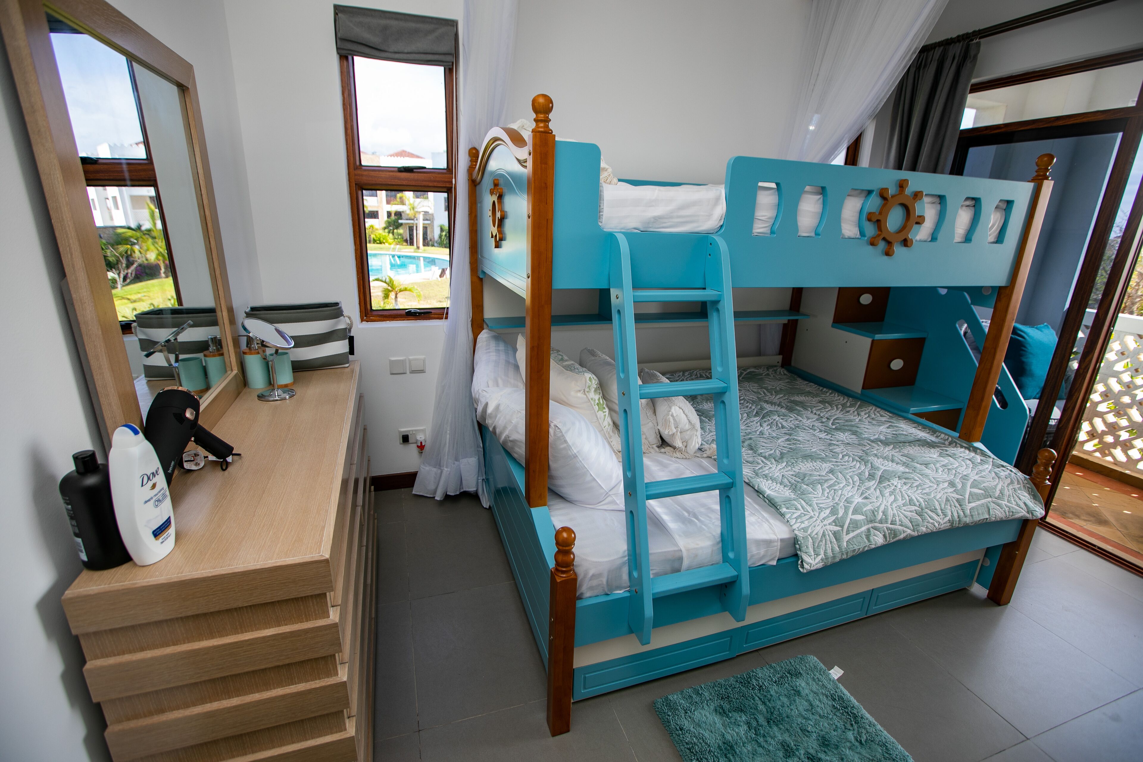 A bunk bed with a wooden frame and a blue mattress, a wooden desk with a mirror, and a window with a view of a pool.