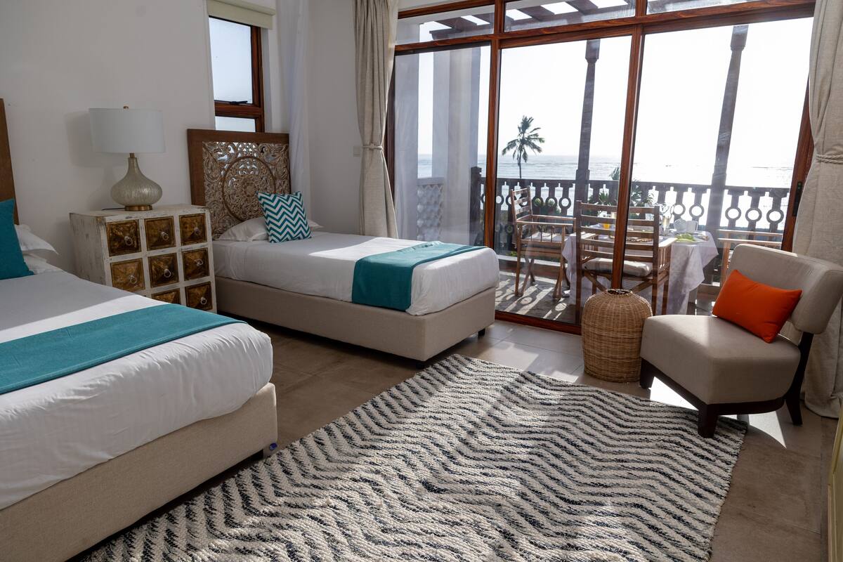 Habitación de hotel con dos camas, un balcón con vista al océano y un área de comedor con mesa y sillas.