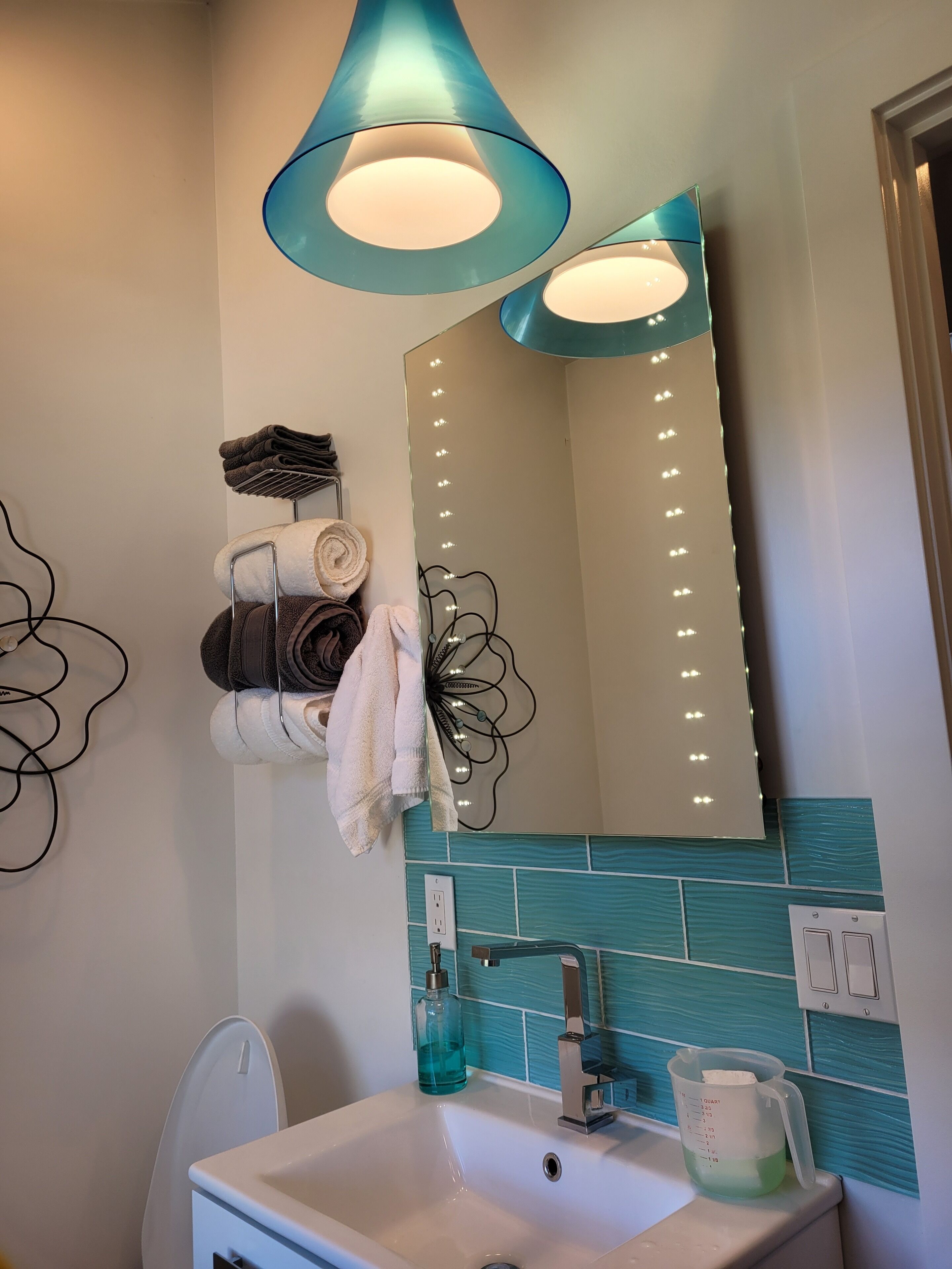 Une salle de bain avec un lavabo blanc, un miroir avec éclairage intégré, un porte-serviettes avec des serviettes et un pare-éclaboussures bleu.