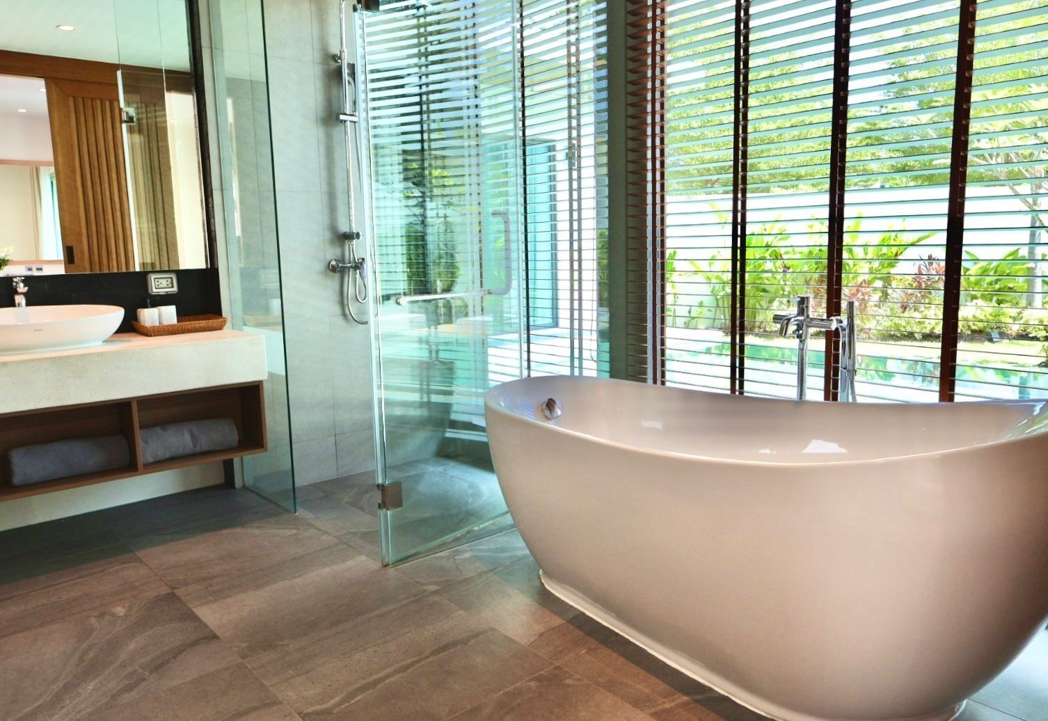 A modern bathroom with a freestanding bathtub, a glass shower enclosure, and a large window with blinds.