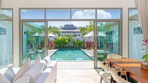 Zona de piscina moderna al aire libre con vista a un edificio tradicional en el fondo, rodeada de exuberante vegetación y equipada con una mesa de madera y sillas blancas.