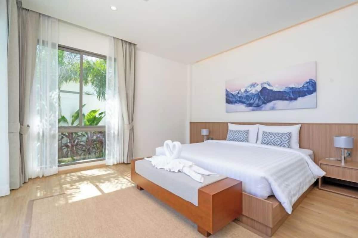 A hotel room with a bed, a bedside table, a nightstand, a window with curtains, and a painting on the wall.