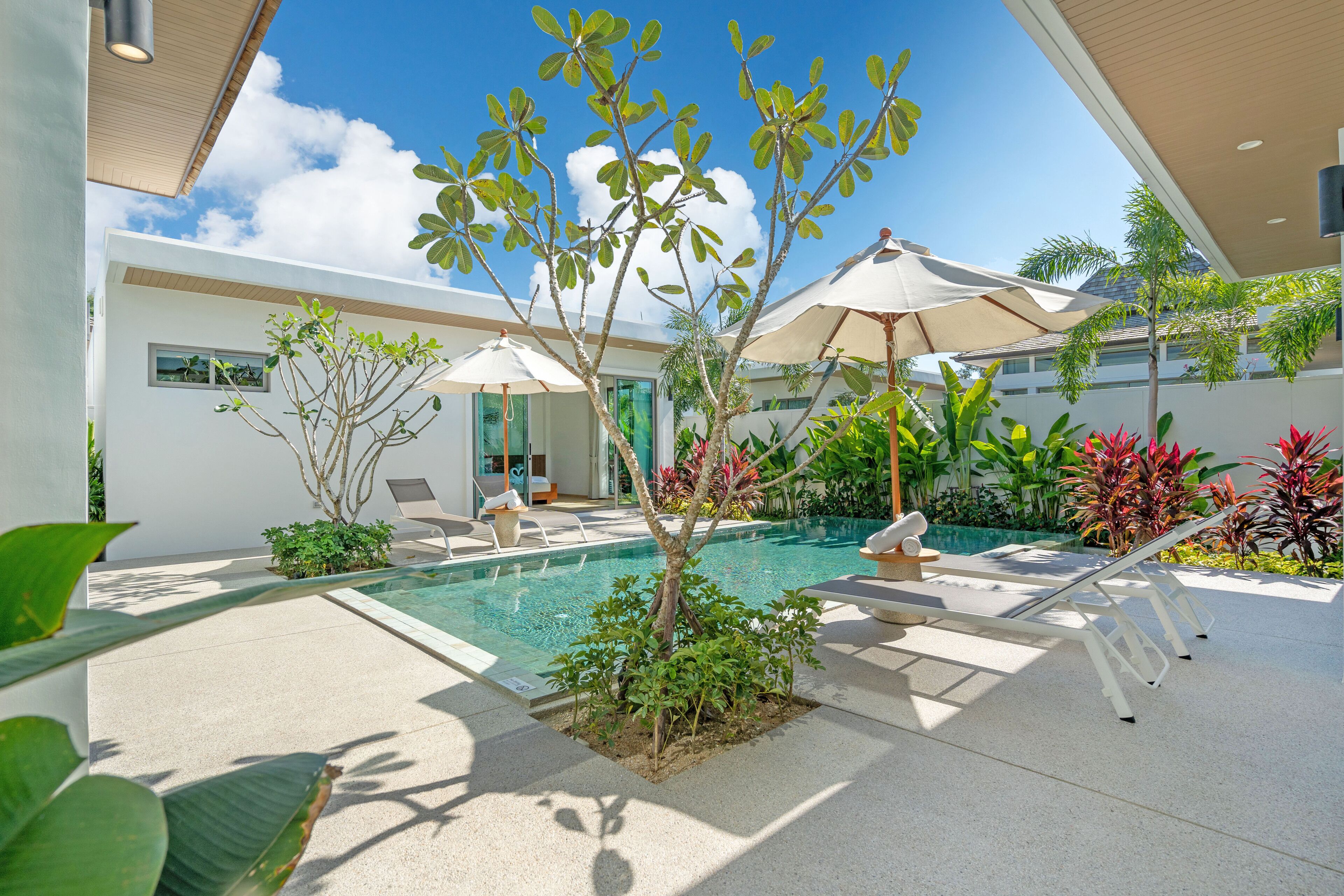 A modern outdoor pool area with lounge chairs and umbrellas, surrounded by greenery and a clear blue sky.