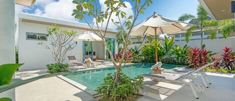 A modern outdoor pool area with lounge chairs and umbrellas, surrounded by greenery and a clear blue sky.