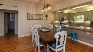 A dining area with a wooden table and white chairs, a kitchen with white cabinets, and a 'Beachhouse' sign on the wall.