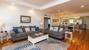 A spacious living area with two grey sofas, a coffee table, and a patterned rug. A kitchen with wooden cabinets and a dining area with a white table and chairs is visible in the background.