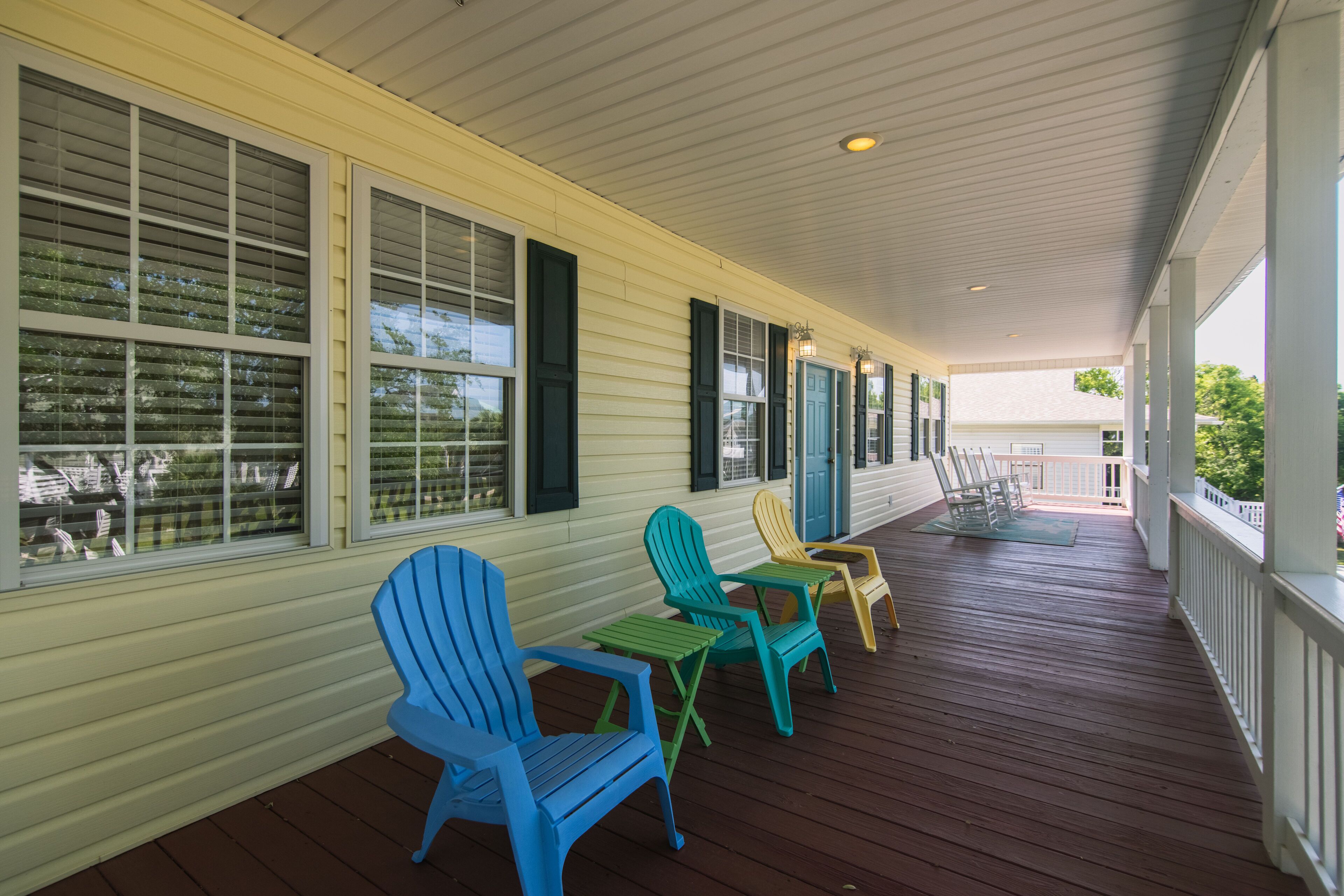 A covered porch with wooden flooring, a row of colorful Adirondack chairs, and large windows with shutters.