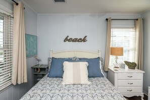 Een slaapkamer met een bed, nachtkastje, lamp en een muur met een bord waarop 'beach' staat.