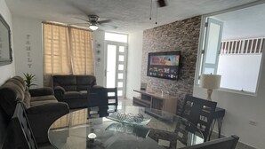 A modern living room with a glass dining table, comfortable sofas, a stone accent wall, a ceiling fan, and a TV mounted on the wall. - Lucor D- Eight House (Rincon PR)