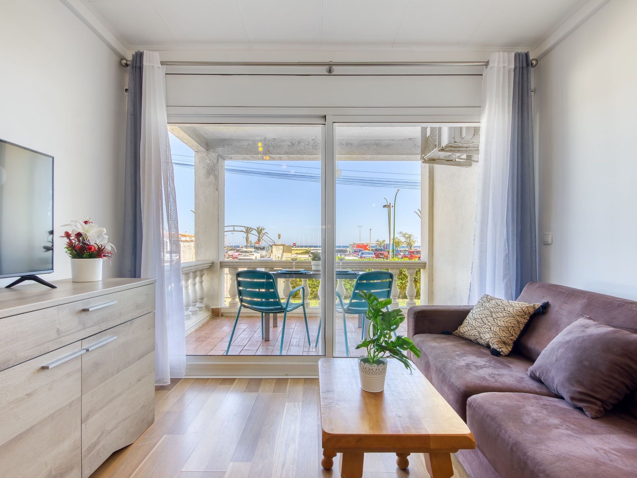 A modern living room with a sofa, a coffee table, and a TV on a cabinet. There is a balcony with a view of the street and parked cars.