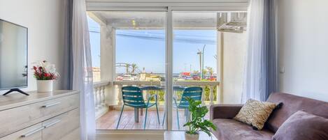 Een moderne woonkamer met een bank, een salontafel en een tv op een kast. Er is een balkon met uitzicht op de straat en geparkeerde auto's.