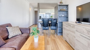 A modern living room with a brown sofa, a wooden coffee table, and a potted plant. There is a kitchen area with a dining table and chairs, and a TV on a wooden cabinet.