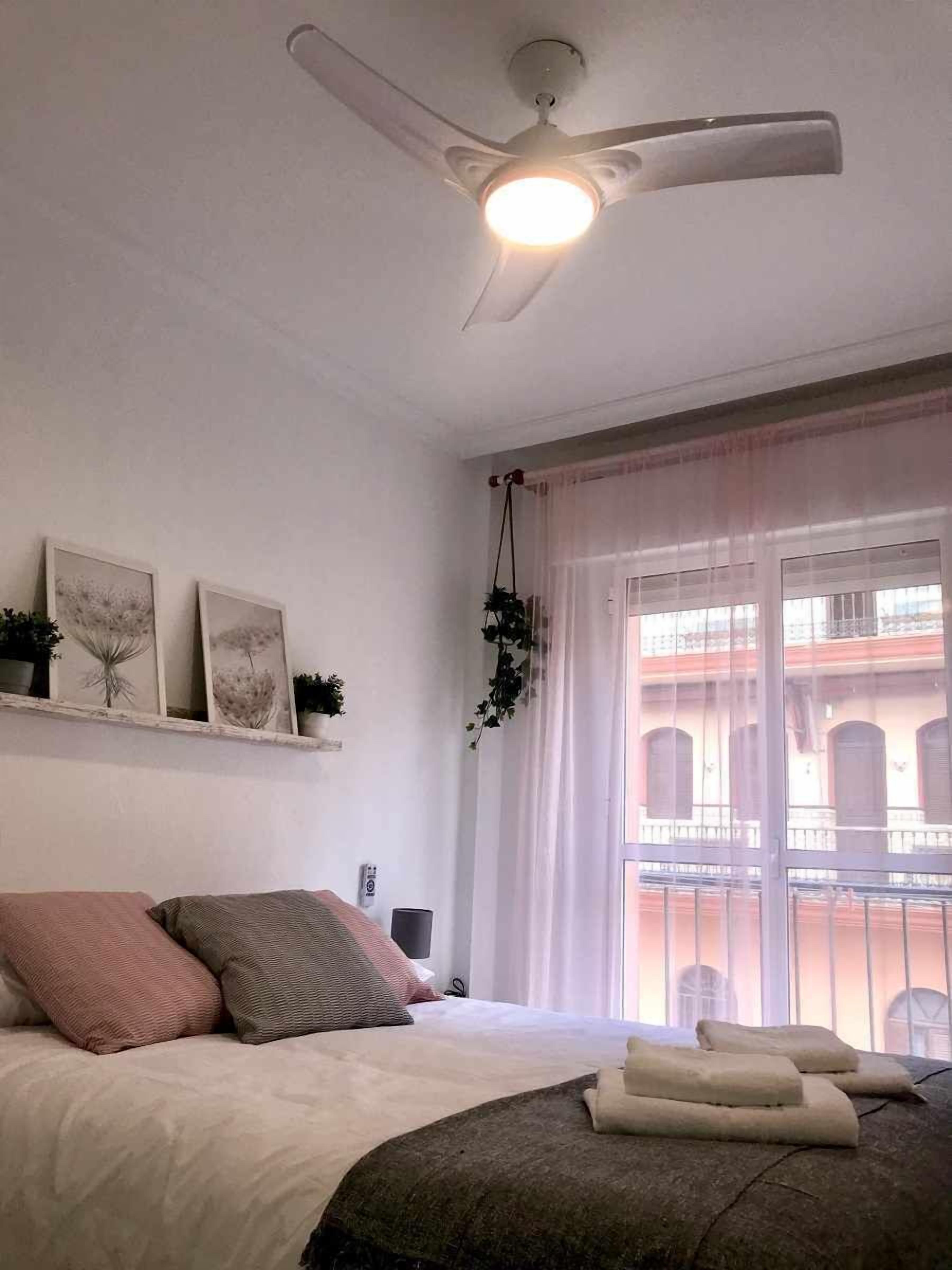Un dormitorio con cama, ventilador de techo, ventana con cortinas e imágenes colgadas en la pared.