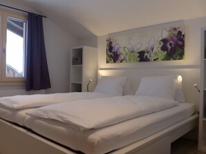 A double bed with white linens in a room with a large floral painting on the wall, a window with curtains, and a white bedside table. - Schwendi 3 by Interhome (Grindelwald)