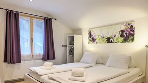 Une chambre moderne avec deux lits, une grande fenêtre ornée de rideaux violets, un radiateur et une œuvre d’art accrochée au mur.