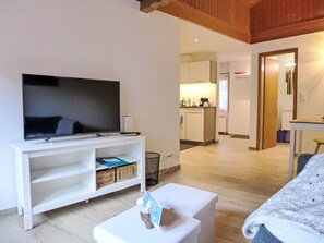 A modern living room with a flat-screen TV on a white stand, a small white table, and a sofa. A kitchen is visible in the background with white cabinets and a sink. - Schwendi 3 by Interhome (Grindelwald)
