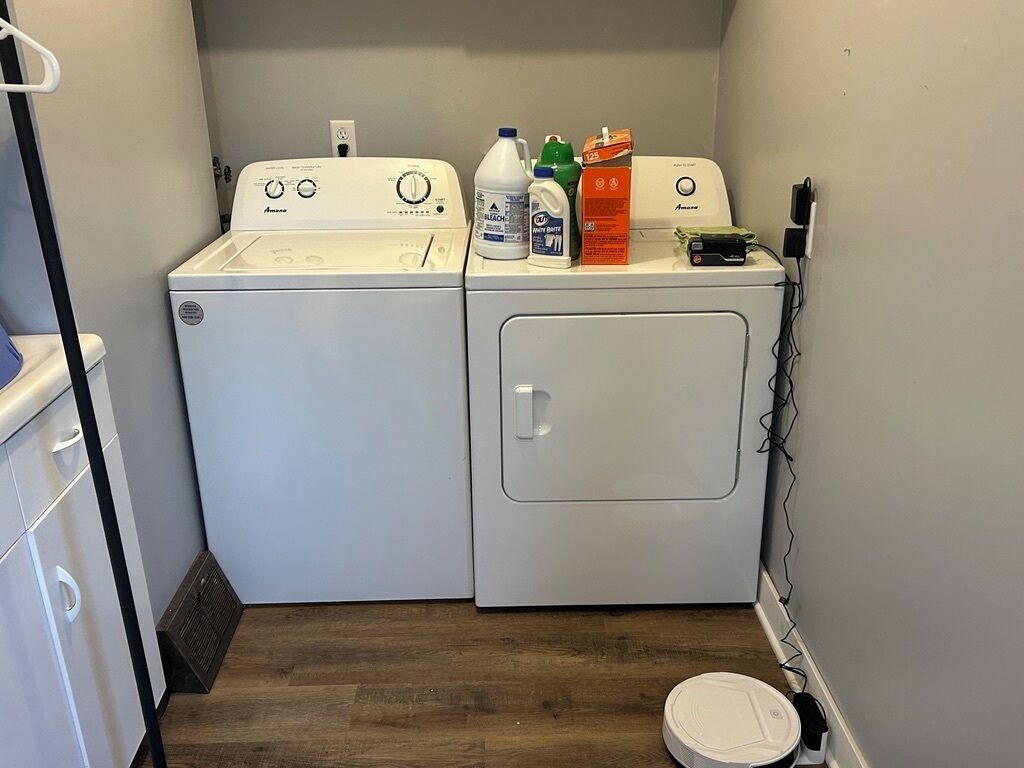 A laundry room with a front-loading washer and dryer, a wooden floor, and various cleaning supplies on the dryer.