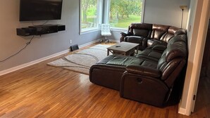 A living room with a leather sofa, a flat-screen TV mounted on the wall, and large windows offering a view of the outdoors.