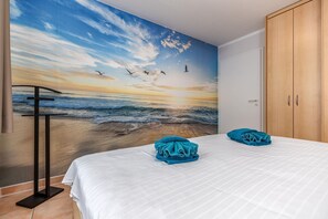 A bedroom with a mural of a beach at sunset, a bed with white bedding, and two blue towels.