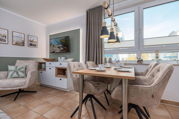 Ein modernes Wohnzimmer mit Essbereich, ausgestattet mit einem Holztisch, gemütlicher Sitzgelegenheit, einem an der Wand montierten Fernseher und einem großen Fenster mit Vorhängen.