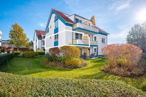 A modern two-story house with a balcony, surrounded by well-maintained gardens and a lawn.