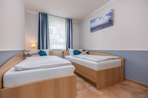 Hotellihuone, jossa on kaksi puista sänkyä, sinisillä verhoilla varustettu ikkuna, seinällä taulu ja yöpöydällä lamppu.