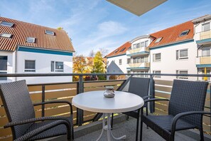 A balcony with wicker chairs and a small table, overlooking a row of buildings with red-tiled roofs.