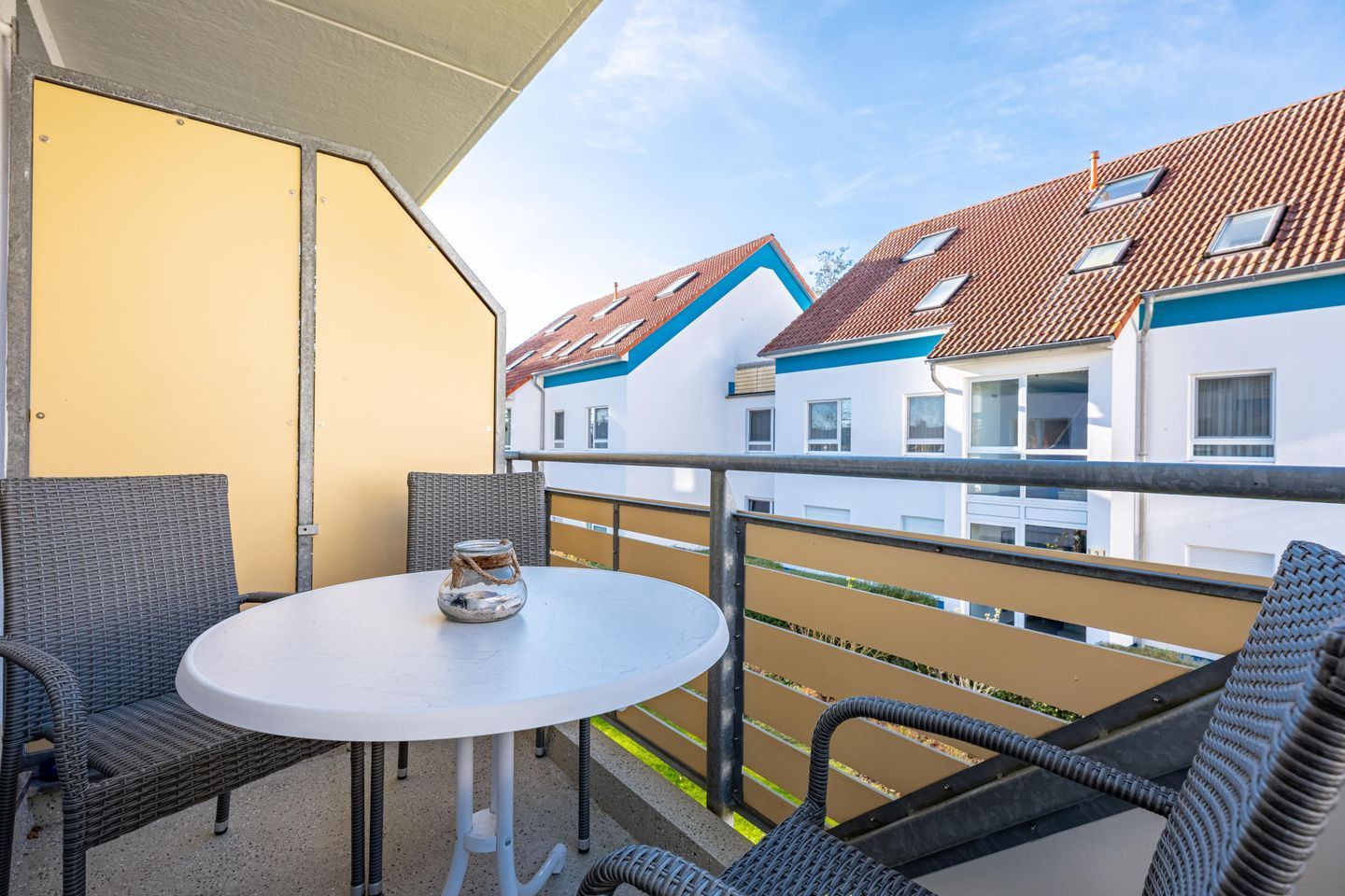 Un balcon avec des fauteuils en osier, une petite table ronde et une vue sur les immeubles voisins.
