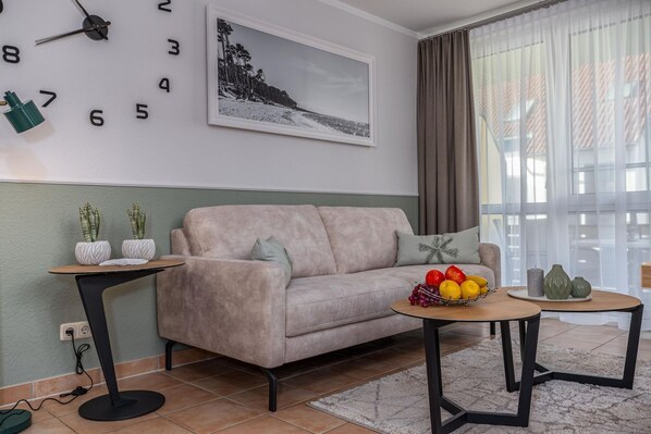 A living room with a sofa, two side tables, a fruit bowl, a wall clock, and a framed picture.