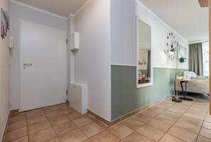 A hotel room with a door, a small cabinet, a mirror, a clock, a bed, a side table, and a window with curtains.