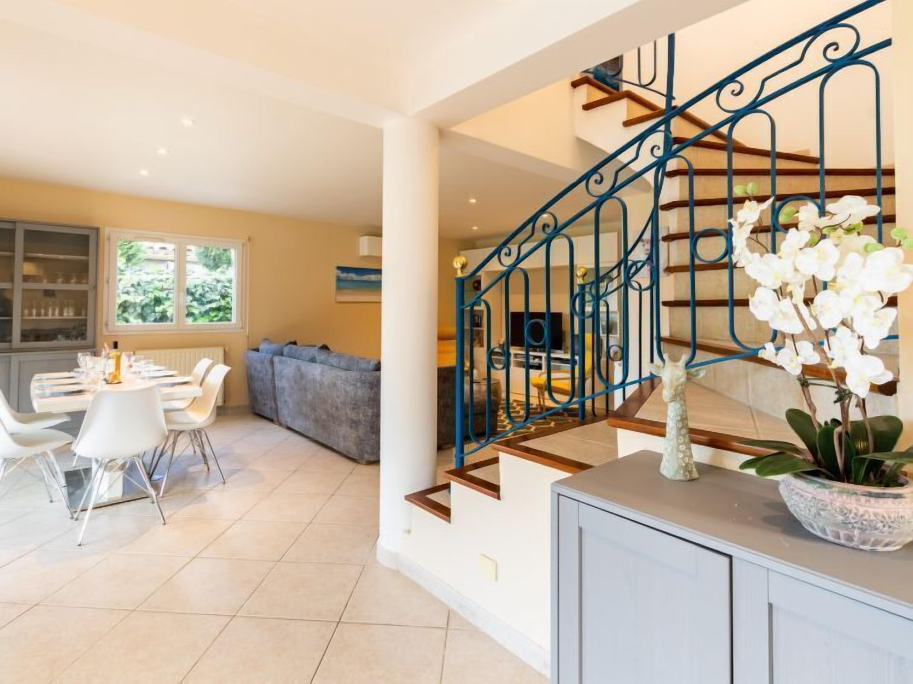 A modern living room with a dining area, a staircase with blue railings, and a view of the outdoors through large windows.