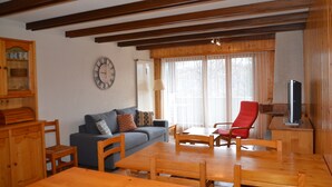 A cozy living room with a wooden dining table, a sofa, and a red chair. A television is on a wooden stand, and a clock is on the wall. - Nice flat for rent in les Collons (Thyon-Les Collons)
