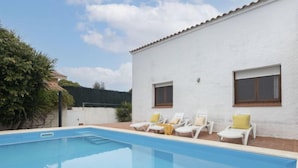 Une piscine bordée de chaises longues blanches et de coussins jaunes, un bâtiment blanc doté de deux fenêtres et un ciel d’un bleu limpide.