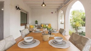 Un coin repas avec une table en bois dressée pour quatre personnes, entourée de chaises rembourrées. On y trouve deux lampes fixées au mur, une fenêtre donnant sur un paysage verdoyant et une porte en arc menant à l’extérieur.