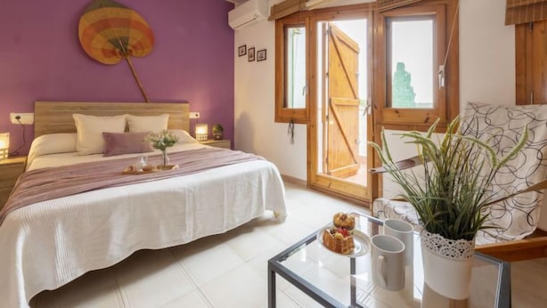 Une chambre à coucher avec un lit, des tables de chevet, une fenêtre avec des volets en bois, une plante en pot et une petite table sur laquelle se trouvent un objet décoratif et deux tasses.