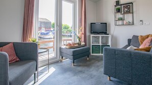 Ein modernes Wohnzimmer mit grauen Sofas, einem Flachbildfernseher und Blick auf Boote durch große Schiebetüren.