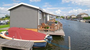 Ein Hausboot liegt an einem Jachthafen, mit einer Deckterrasse, Sitzgelegenheiten im Freien und einem kleinen Garten.